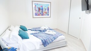 Una cama bien hecha con ropa de cama blanca, mantas estampadas en azul y blanco, y almohadas color turquesa. Un cuadro enmarcado de un paisaje urbano está en la pared sobre la cama.