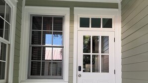 A room with a white door and two windows, one with a view of a building and the other with a clear sky.