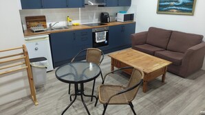 Une cuisine compacte avec des meubles bleus, un coin repas doté d’une table en verre et de chaises en osier, et un canapé marron.