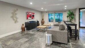 Un salon moderne avec un canapé gris, une table basse et un téléviseur fixé au mur. On y trouve également un coin repas avec une table et des chaises, ainsi qu’une grande fenêtre offrant une vue sur l’extérieur.