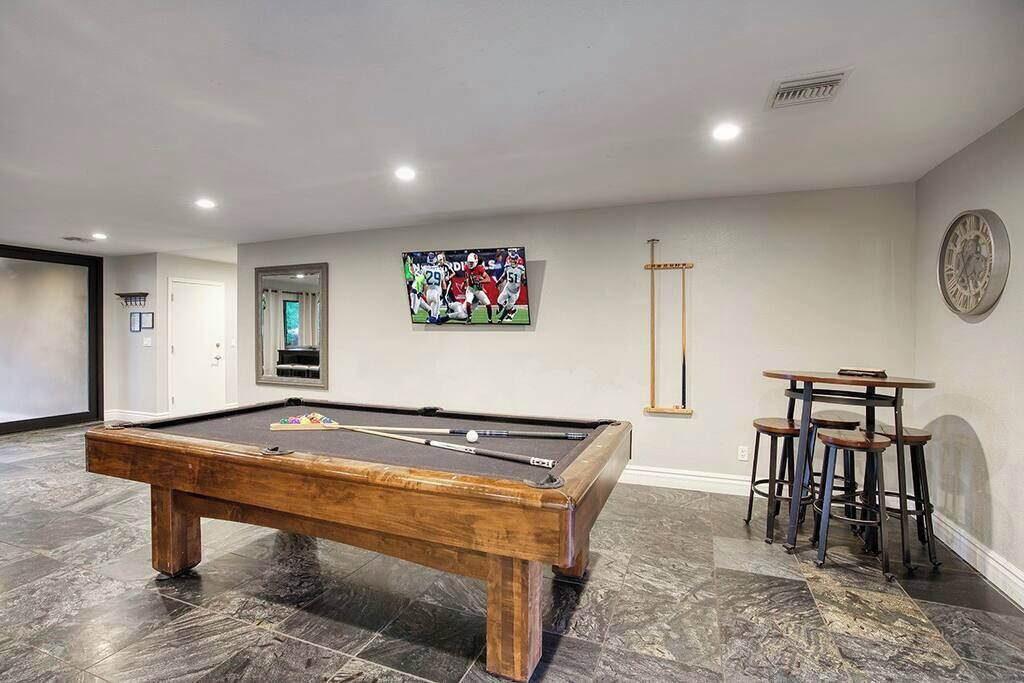Une table de billard dans une pièce dont le sol est carrelé, un coin bar avec des tabourets, une horloge au mur et un tableau encadré représentant une scène sportive.