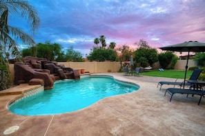 A swimming pool with a rock feature, surrounded by a patio with lounge chairs and an umbrella.