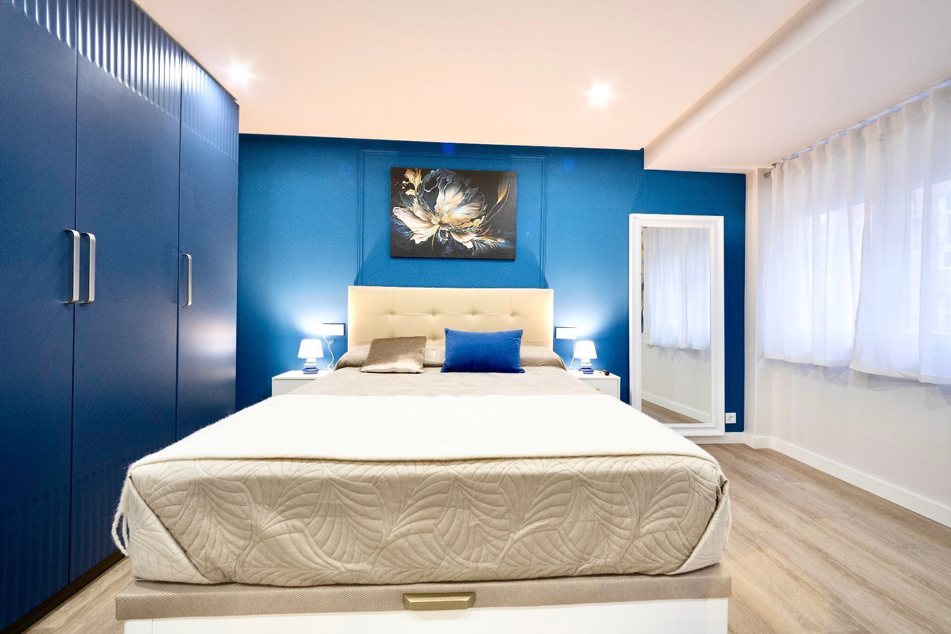 Um quarto moderno com uma cama grande, uma cabeceira, abajures e uma parede azul com um quadro floral.