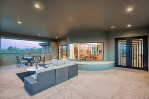 An outdoor living area with a seating arrangement, a dining area visible through a glass partition, and a view of the landscape. - Scottsdale Skyline Views with Waterfall Patio, Sauna, Steam Room & Games (Paradise Valley)