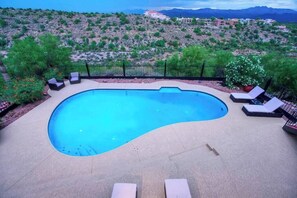 Ein Pool mit geschwungener Kante, umgeben von einer Betonplattform und einem schwarzen Metallzaun. Liegestühle und ein Topfpflanze befinden sich in der Nähe des Pools.
