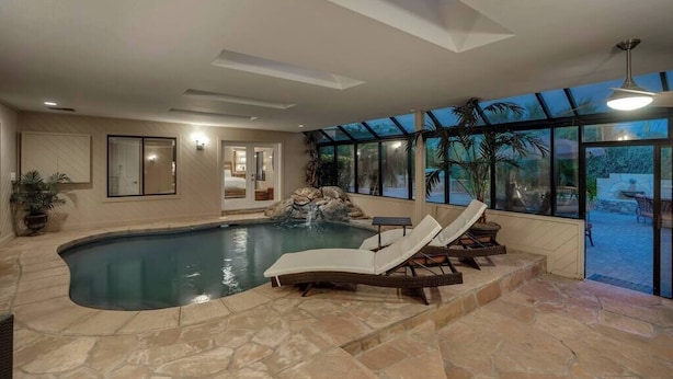 An indoor swimming pool with lounge chairs, a waterfall feature, and large windows offering views of the outdoors. - The Refuge (Fountain Hills)