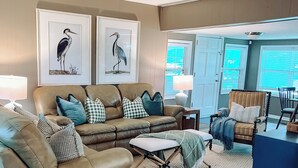 Un soggiorno con un divano angolare in pelle marrone, un tavolino bianco e una poltrona blu. Alle pareti, due quadri incorniciati raffiguranti uccelli; presente anche una ventola a soffitto e una vista all'esterno tramite una porta.