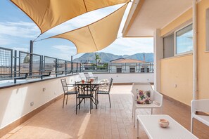 Une terrasse sur le toit, avec une table et des chaises, un grand auvent et une vue sur les bâtiments et les montagnes en arrière-plan.