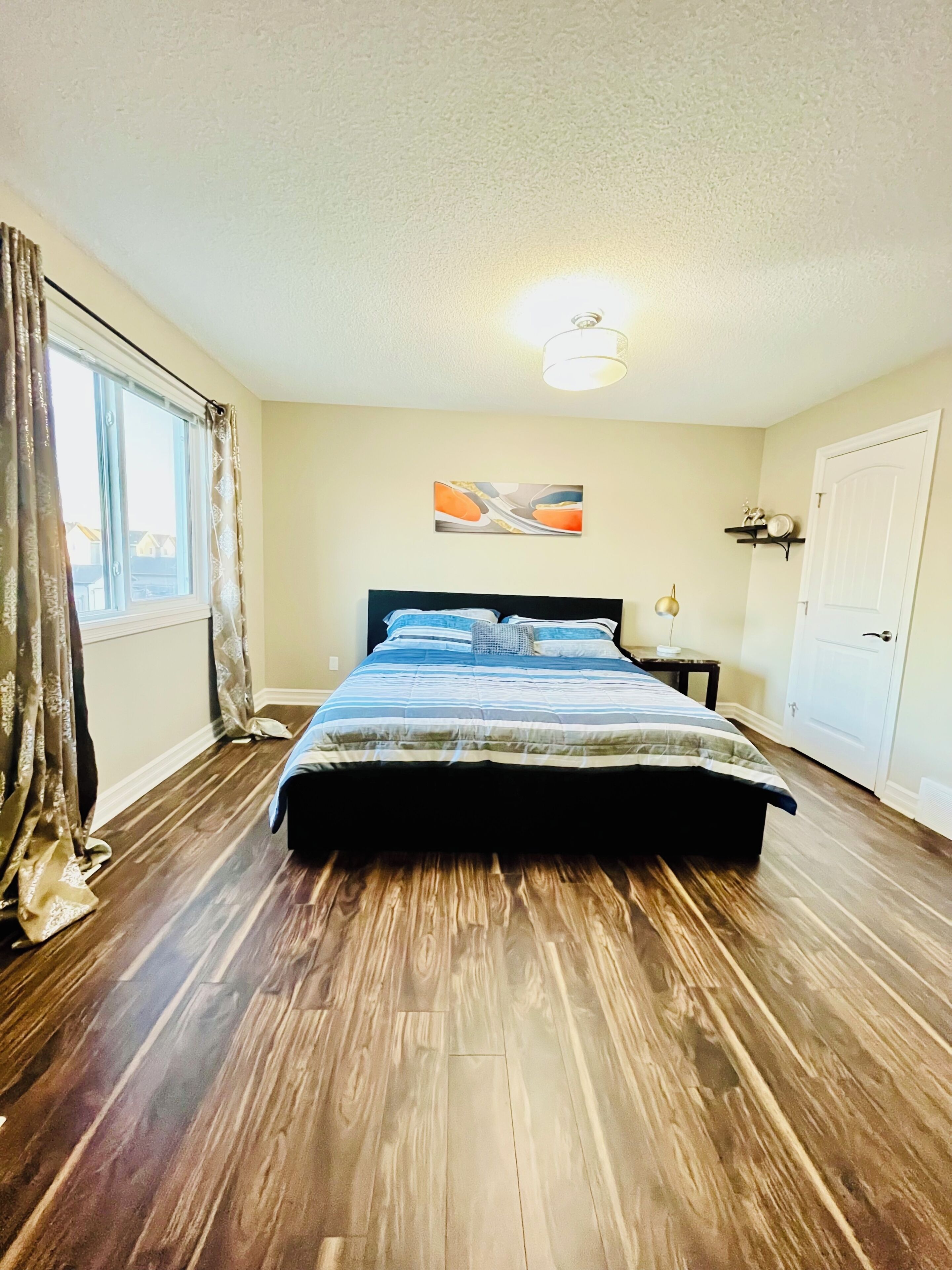 A bedroom with a bed, a nightstand, a wall-mounted shelf, a window with curtains, and a door.