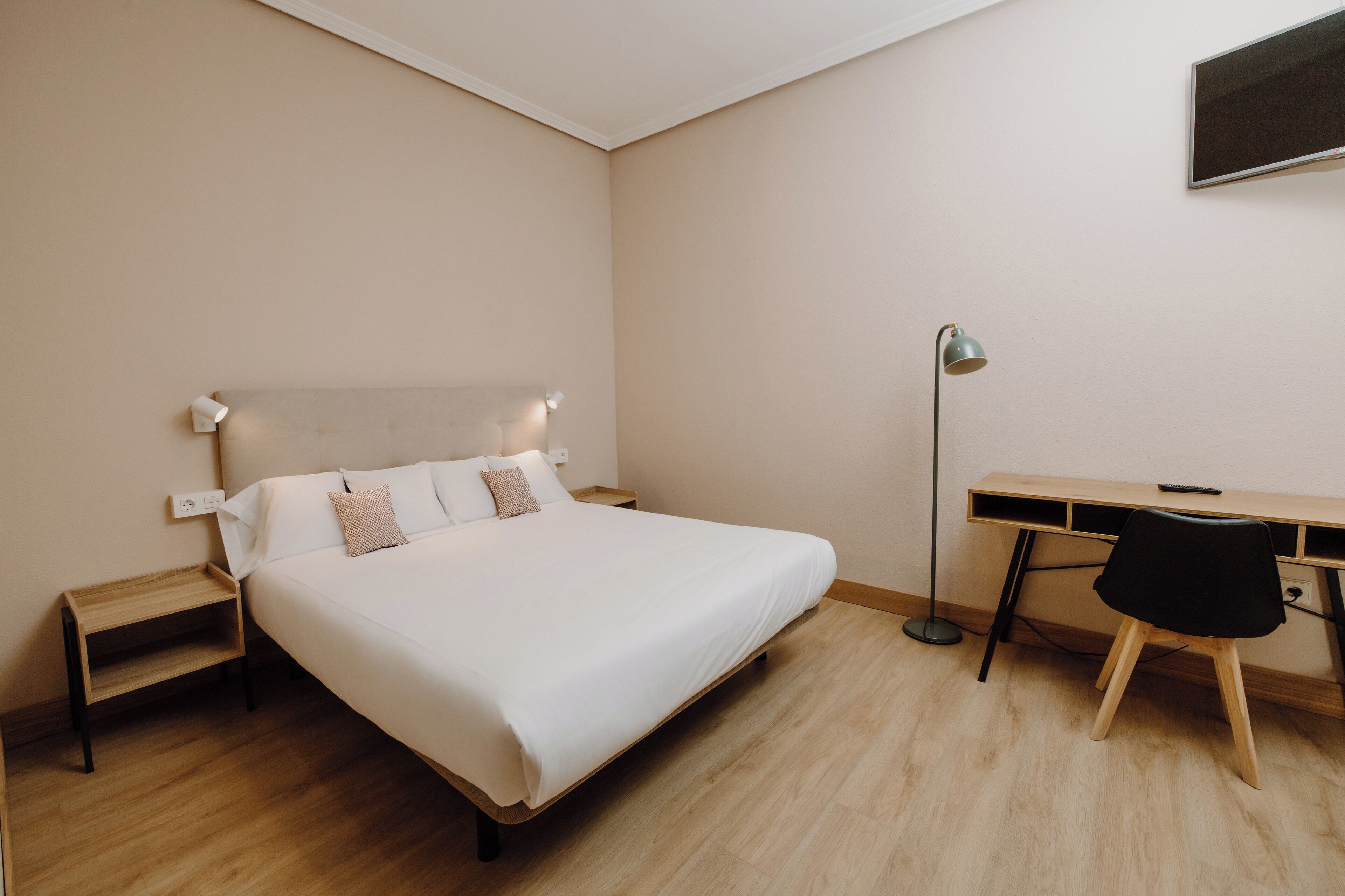 Un dormitorio con cama, mesita de noche, escritorio, silla y televisor en la pared.