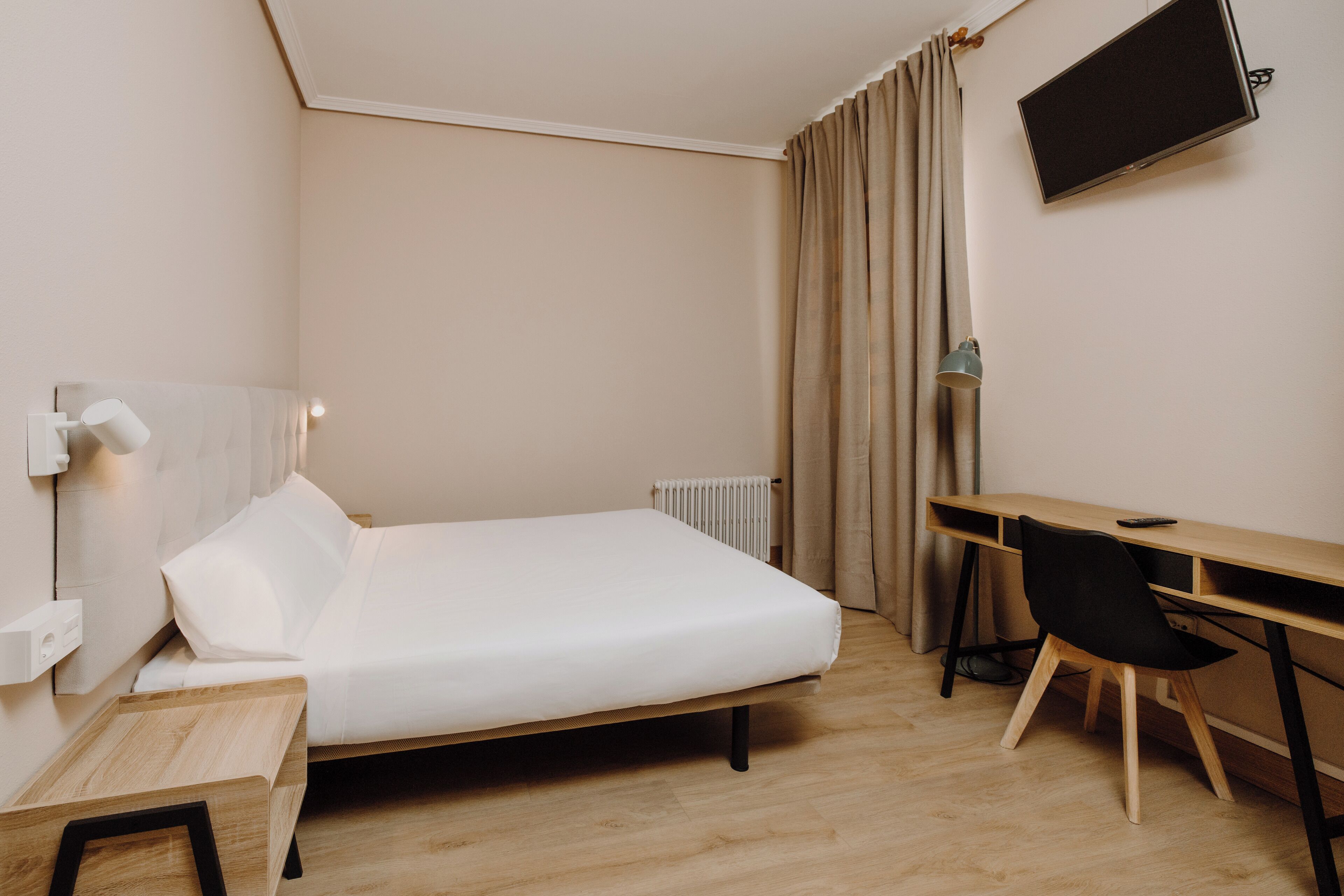 A bedroom with a bed, a desk, a chair, a TV, and a radiator.