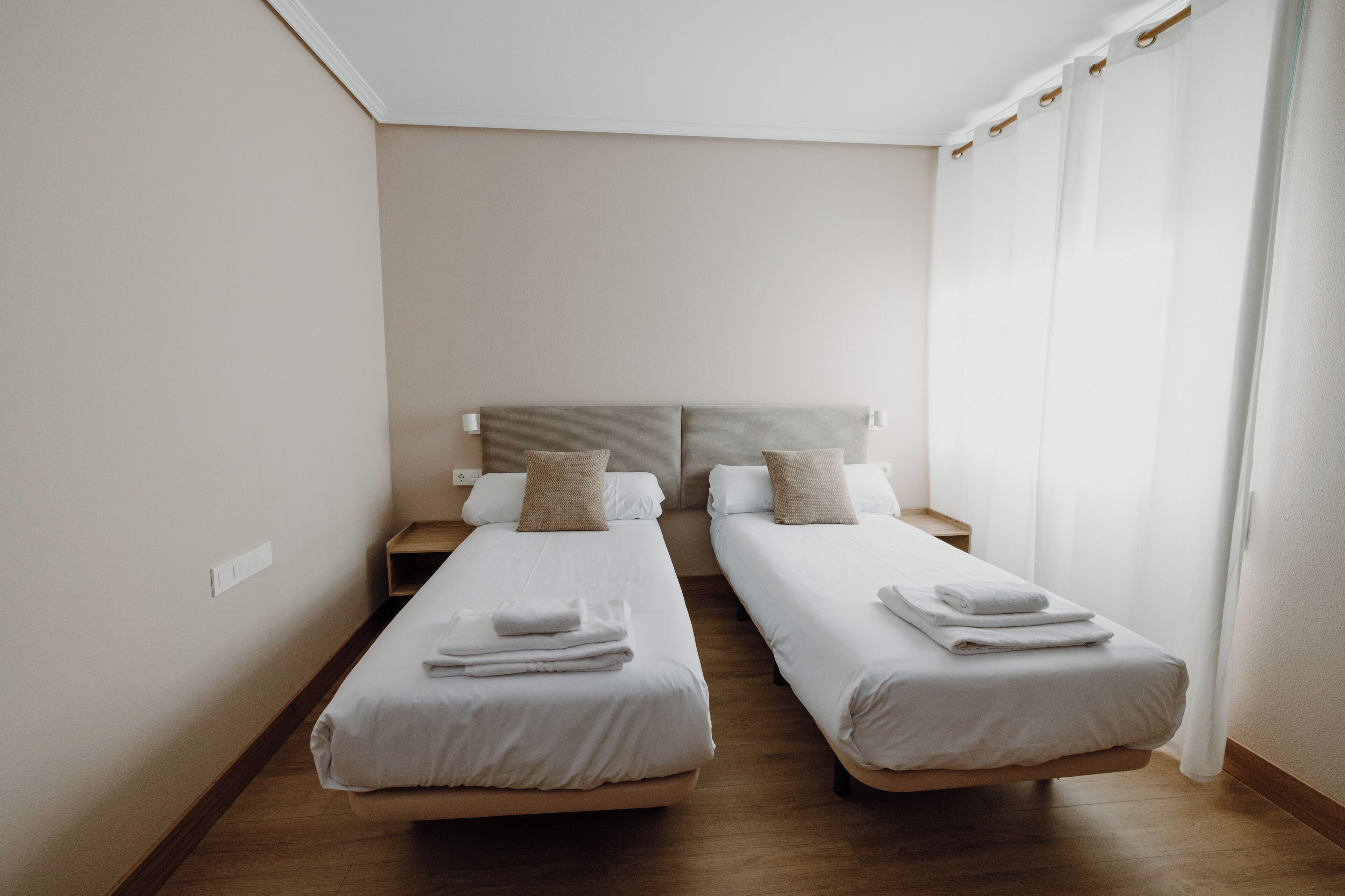 A hotel room with two single beds, each with a wooden headboard, white bedding, and a small bedside table with a lamp.