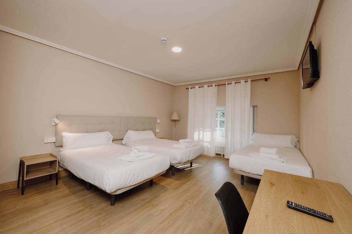 Habitación de hotel con tres camas, una mesa de madera, televisión montada en la pared y una ventana con cortinas.