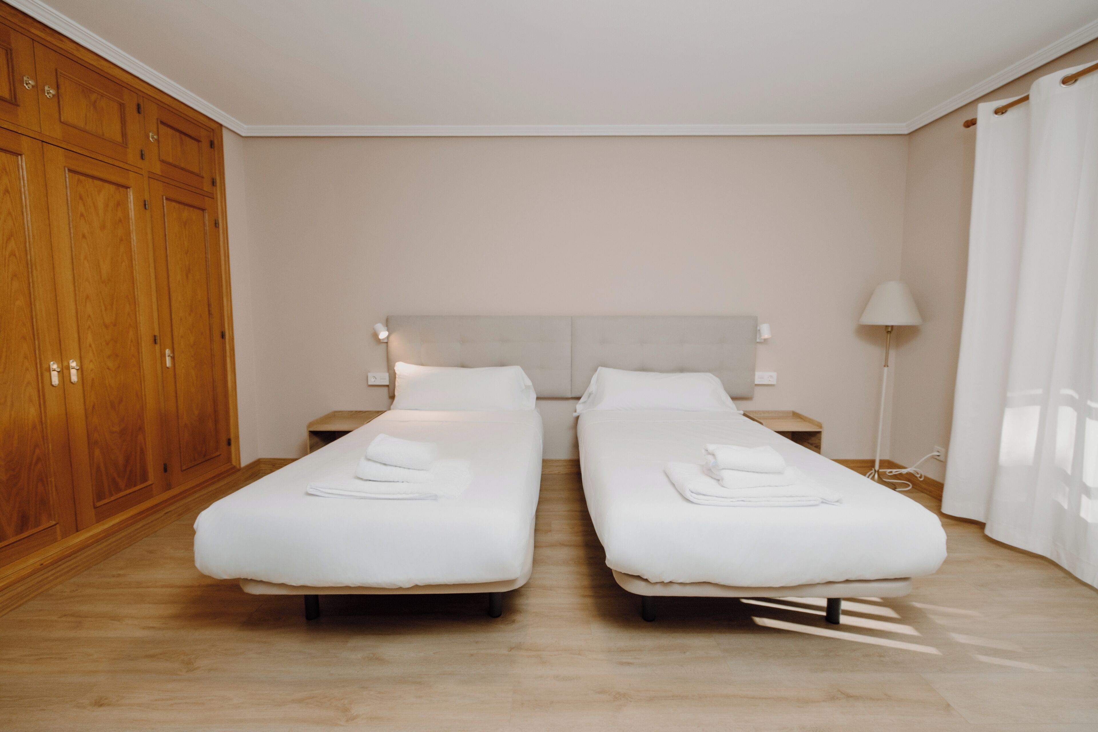 A hotel room with two single beds, wooden wardrobe, bedside tables, and a lamp.
