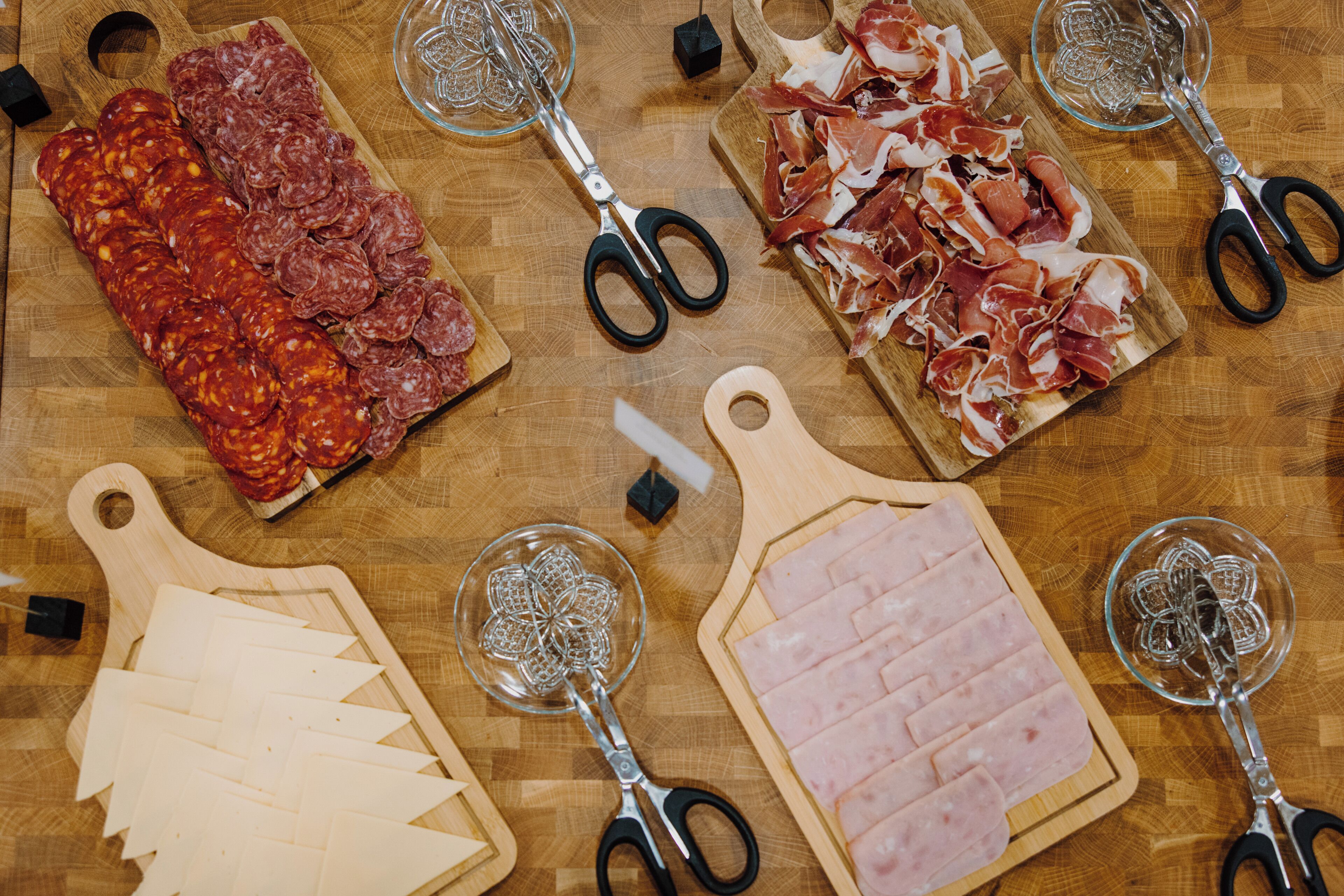 A wooden cutting board with various types of cured meats and cheese, accompanied by a pair of scissors and a glass.