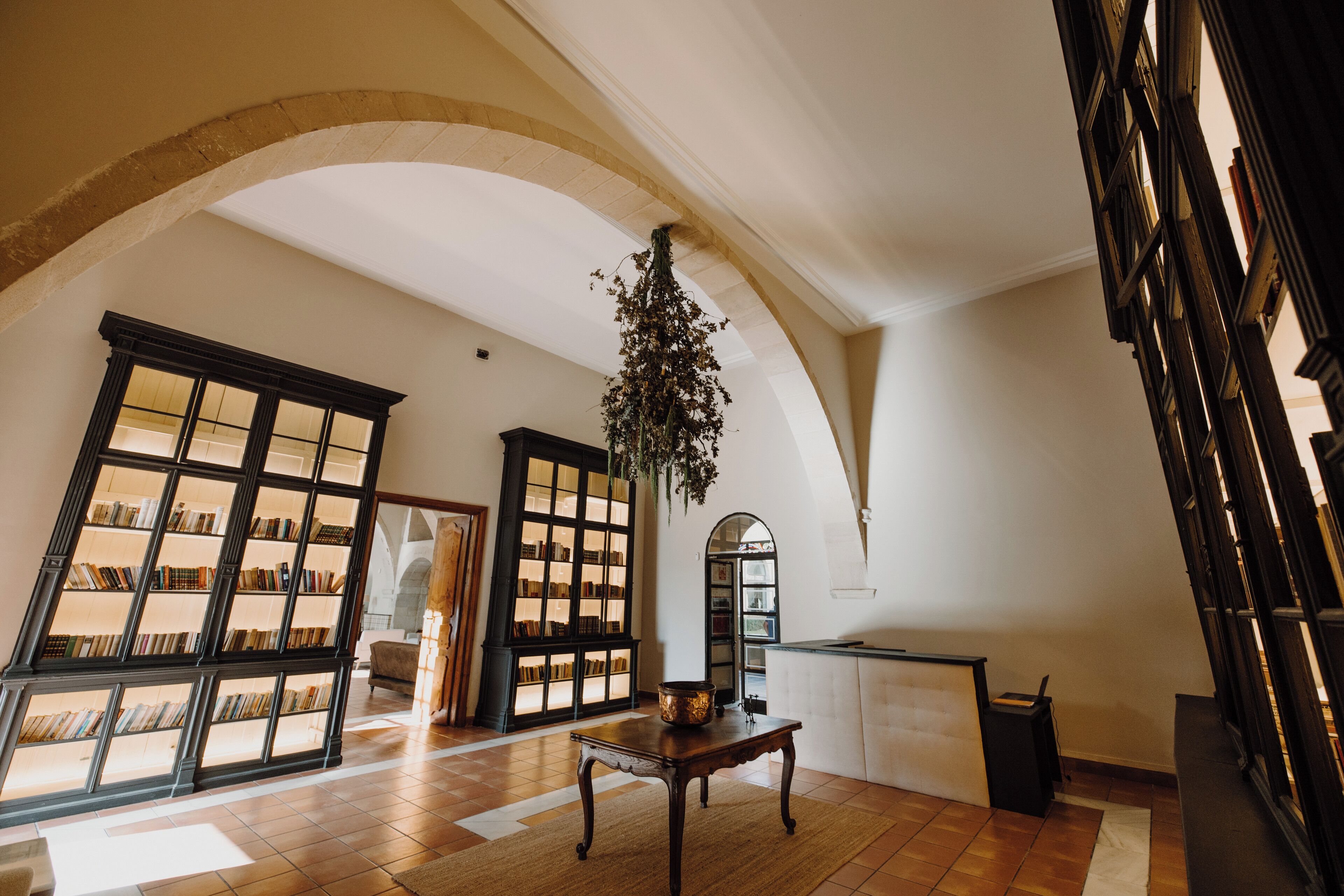 A spacious library with high ceilings and arched windows, featuring bookshelves and a central table.