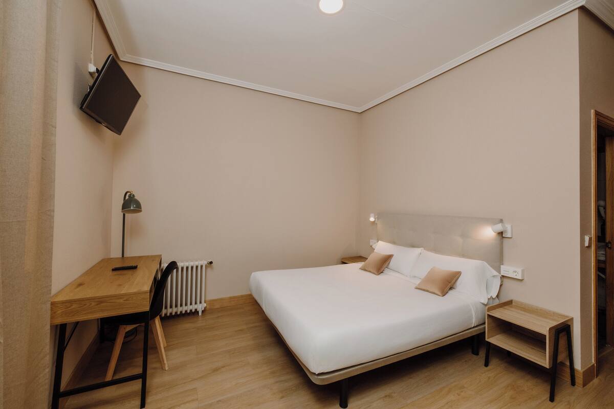 Un dormitorio con cama, mesitas de noche, escritorio, silla, radiador y televisor montado en la pared.