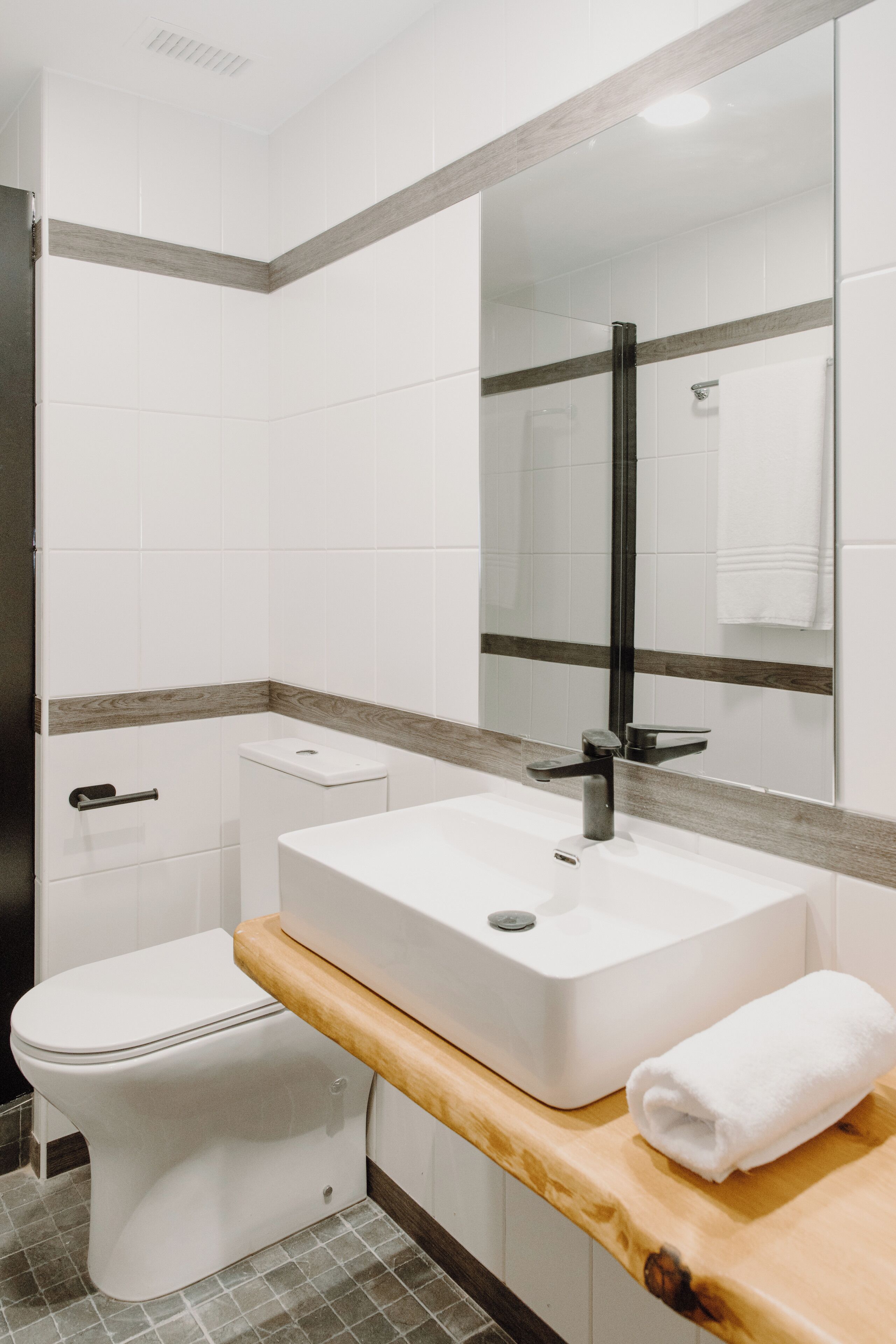 A modern bathroom with a white toilet, a white sink with a wooden countertop, and a large mirror.