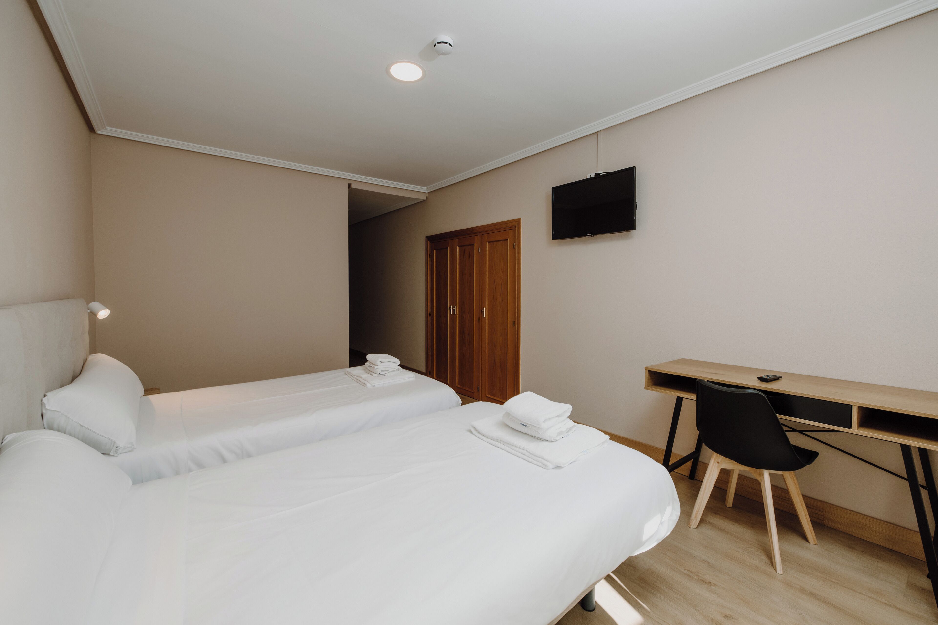 A hotel room with two beds, a desk, a chair, a TV, and a door.