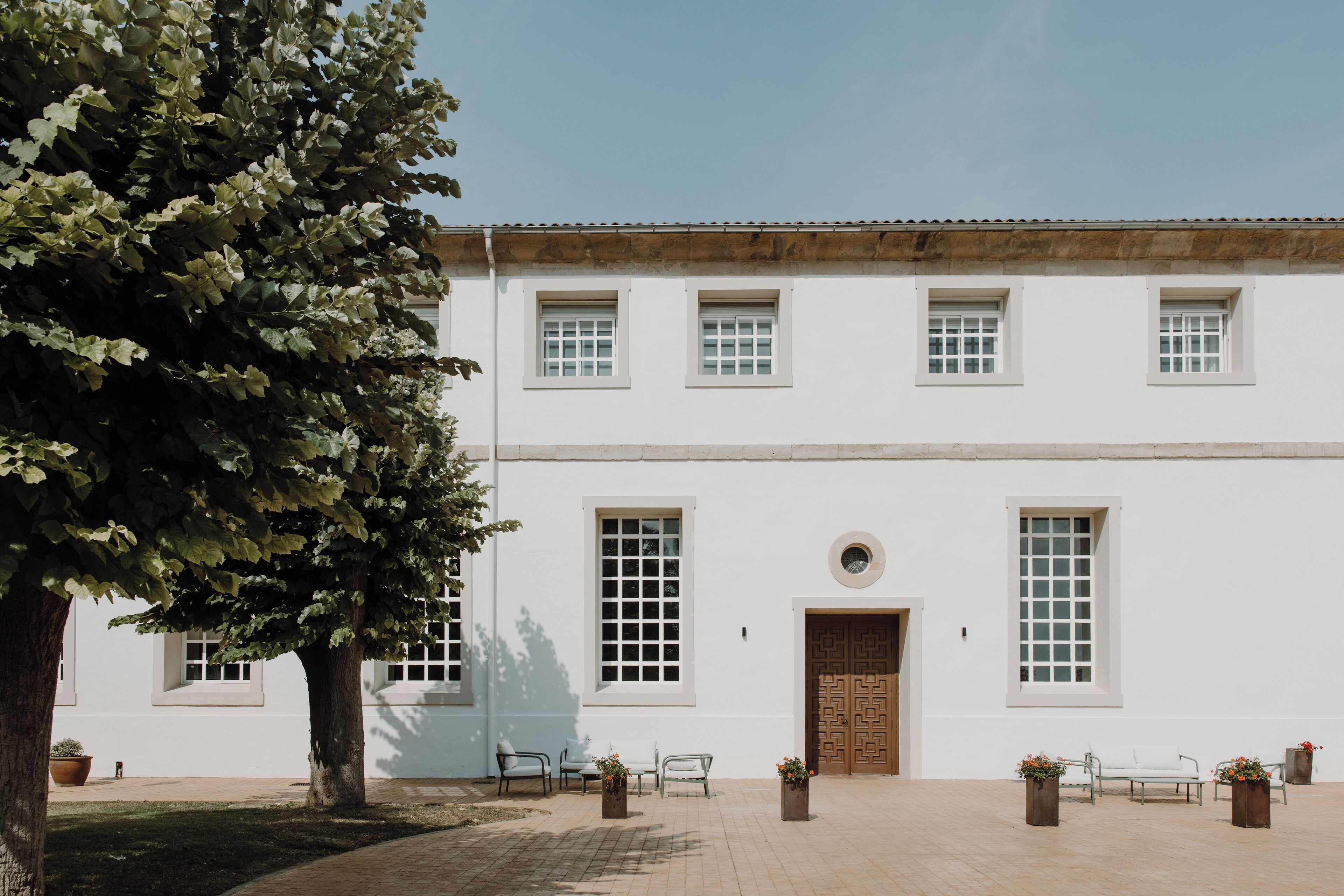 A white building with a tiled roof, large windows, and a central door. There are potted plants and benches in front.