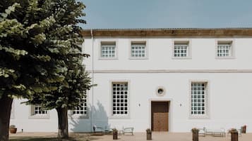 A white building with a tiled roof, large windows, and a central door. There are potted plants and benches in front.