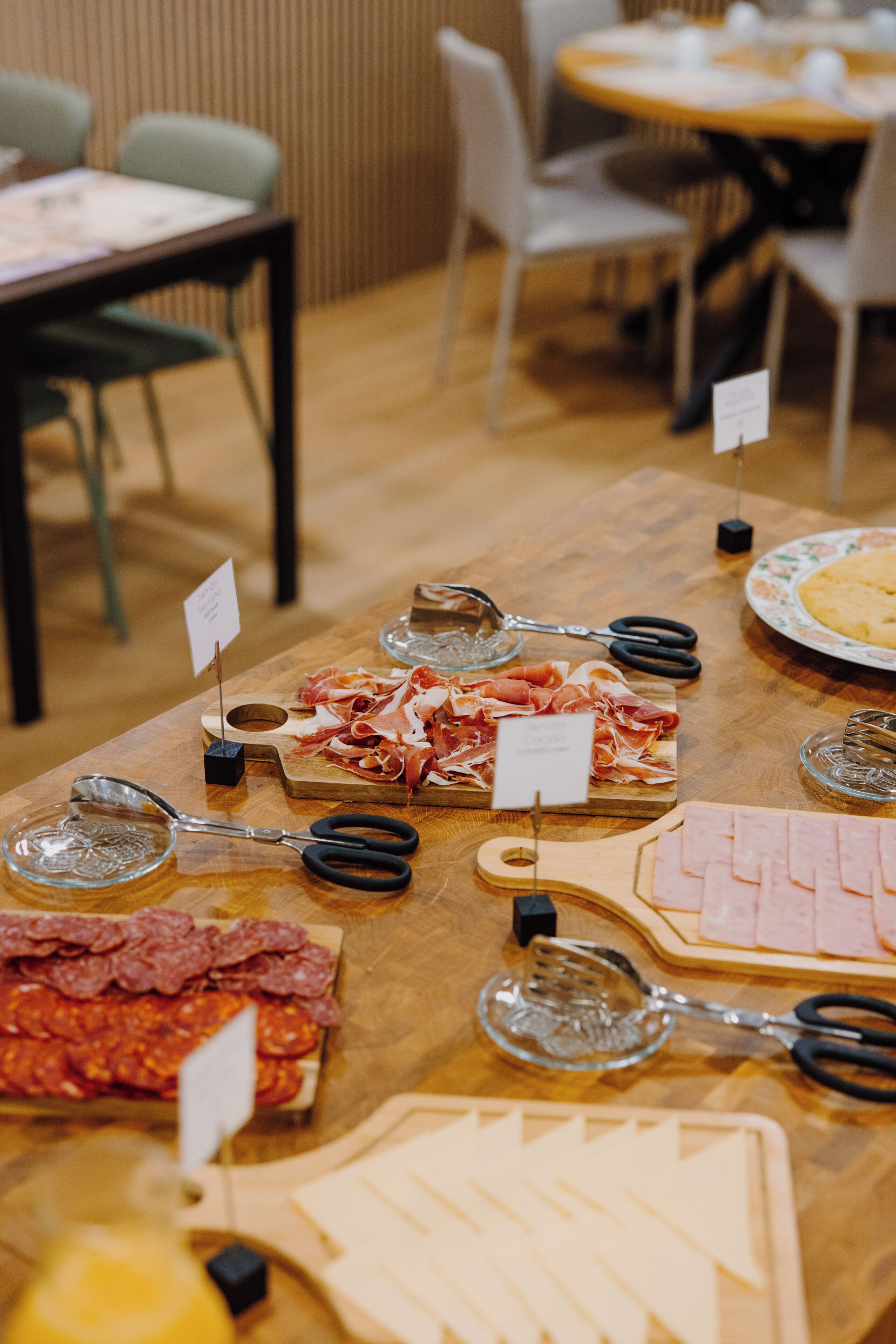 A table with various food items and utensils, including a plate of sliced meats, a glass dish with a dessert, and a pair of scissors.
