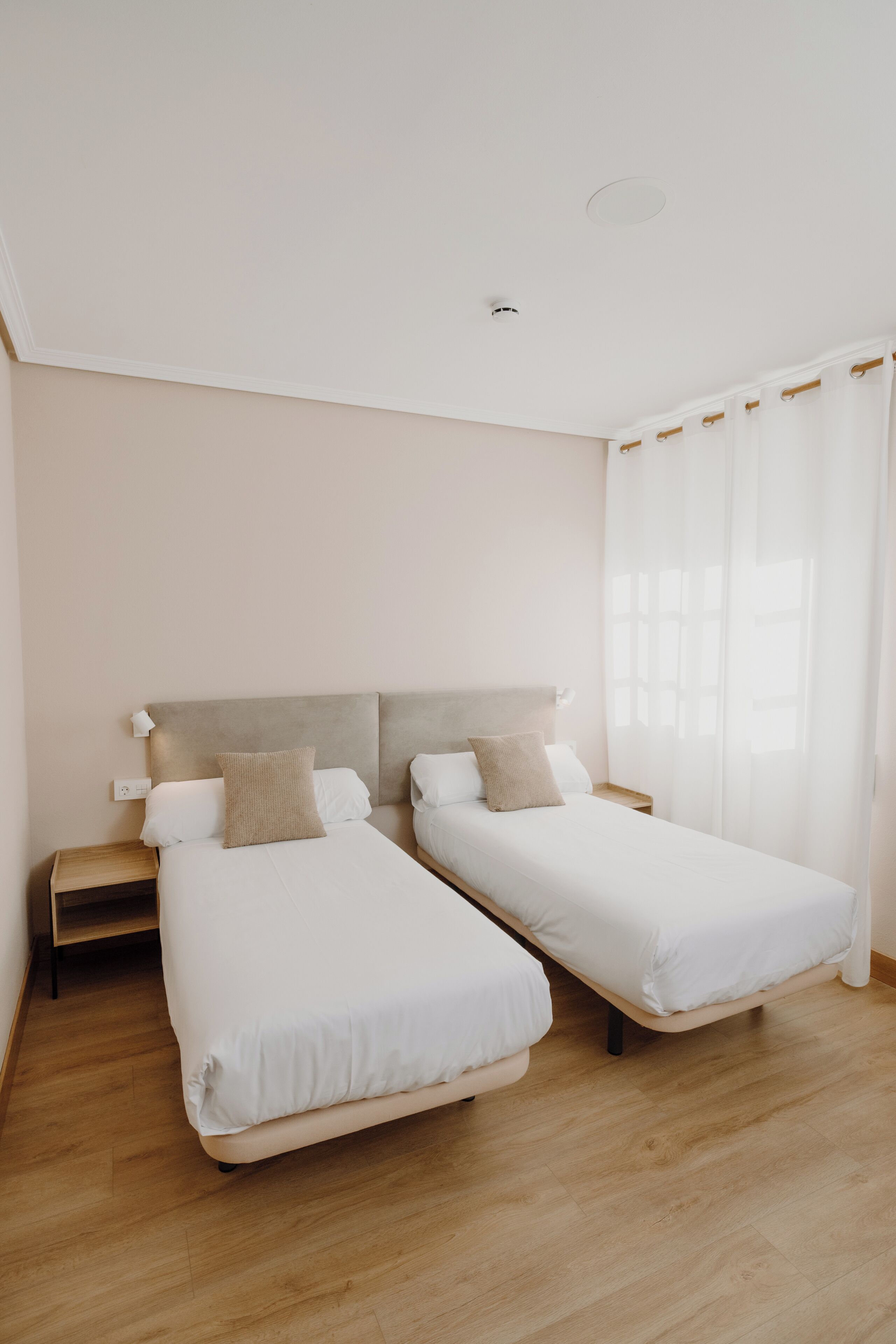 Un dormitorio con dos camas individuales, una mesita de noche de madera, un techo blanco y una ventana con cortinas traslúcidas.