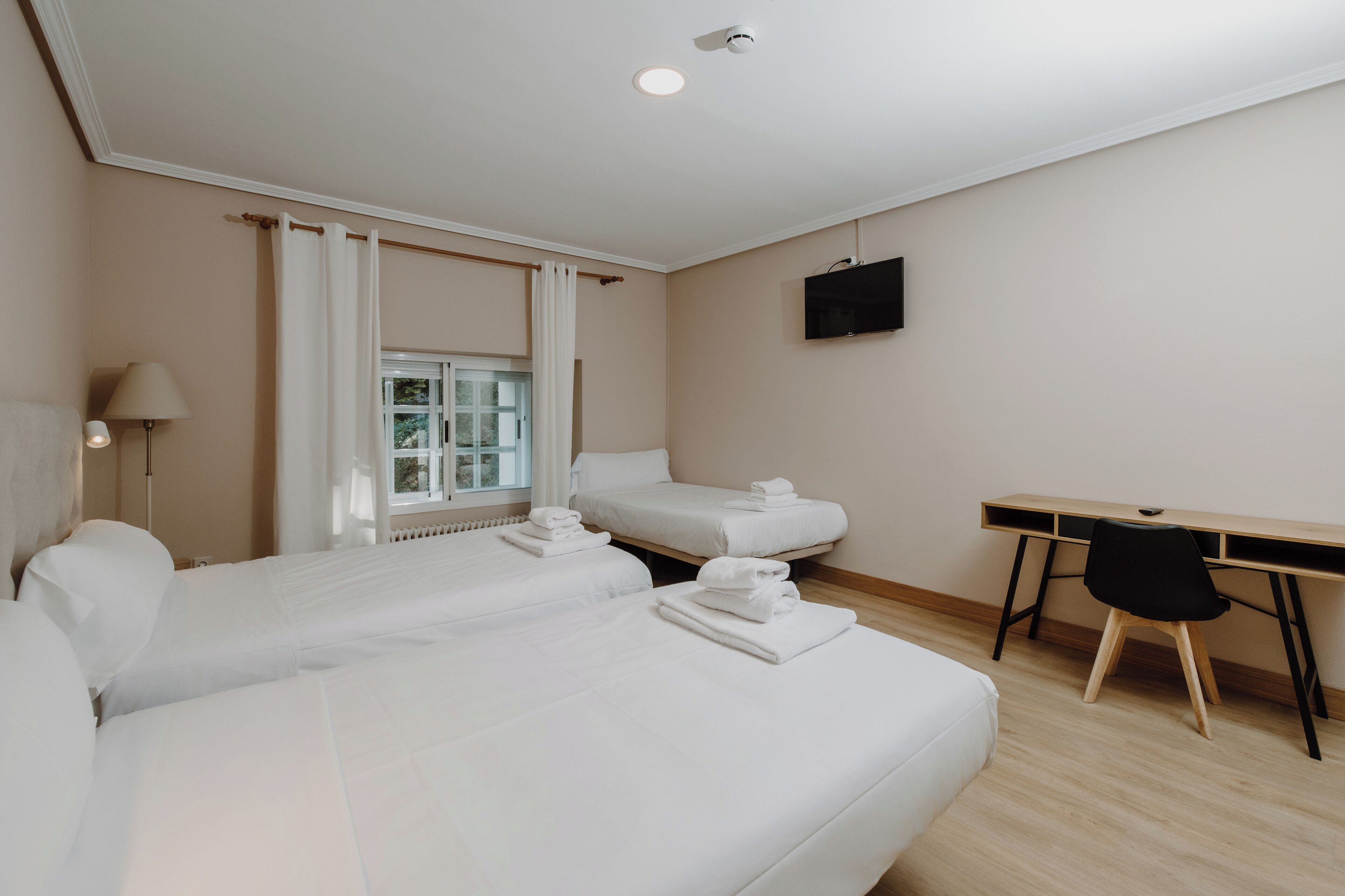 A hotel room with two single beds, a desk, a chair, a TV, and a window with curtains.