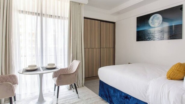A hotel room with a bed, a dining table with chairs, a large window with curtains, and a painting of a moon on the wall.