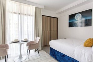 A hotel room with a bed, a dining table with chairs, a large window with curtains, and a painting of a moon on the wall. - Casa Inikas Petite Studio (Nairobi)