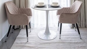 A modern dining area with a round table and two cushioned chairs, a large window with sheer curtains, and a minimalist decor.