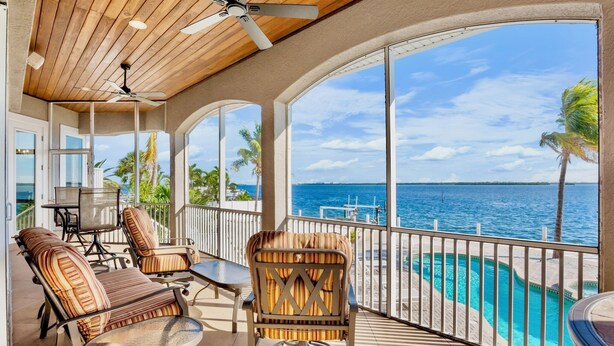 A sunlit balcony with a view of the ocean, furnished with striped chairs and a table, and a ceiling fan.