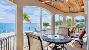 A sunlit patio with a view of the sea, furnished with a round table and chairs, and a ceiling fan.