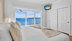 A bedroom with a large window overlooking a body of water, a bed with white bedding, a television mounted on the wall, and a white door.