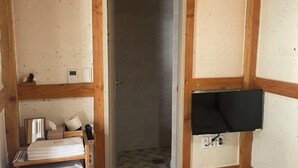 Una habitación pequeña con piso de madera, un televisor montado en la pared, una puerta que da a un baño y un estante con toallas y artículos de aseo.