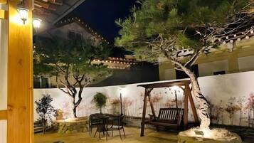 A traditional Japanese-style garden with a wooden pergola, a stone bench, and a pine tree.