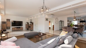 A spacious living area with a large gray sofa, a coffee table, and a TV. There is a dining area with a table and chairs, and a kitchen visible in the background. - Villa Anteo-Split by Villas Guide (Košute)