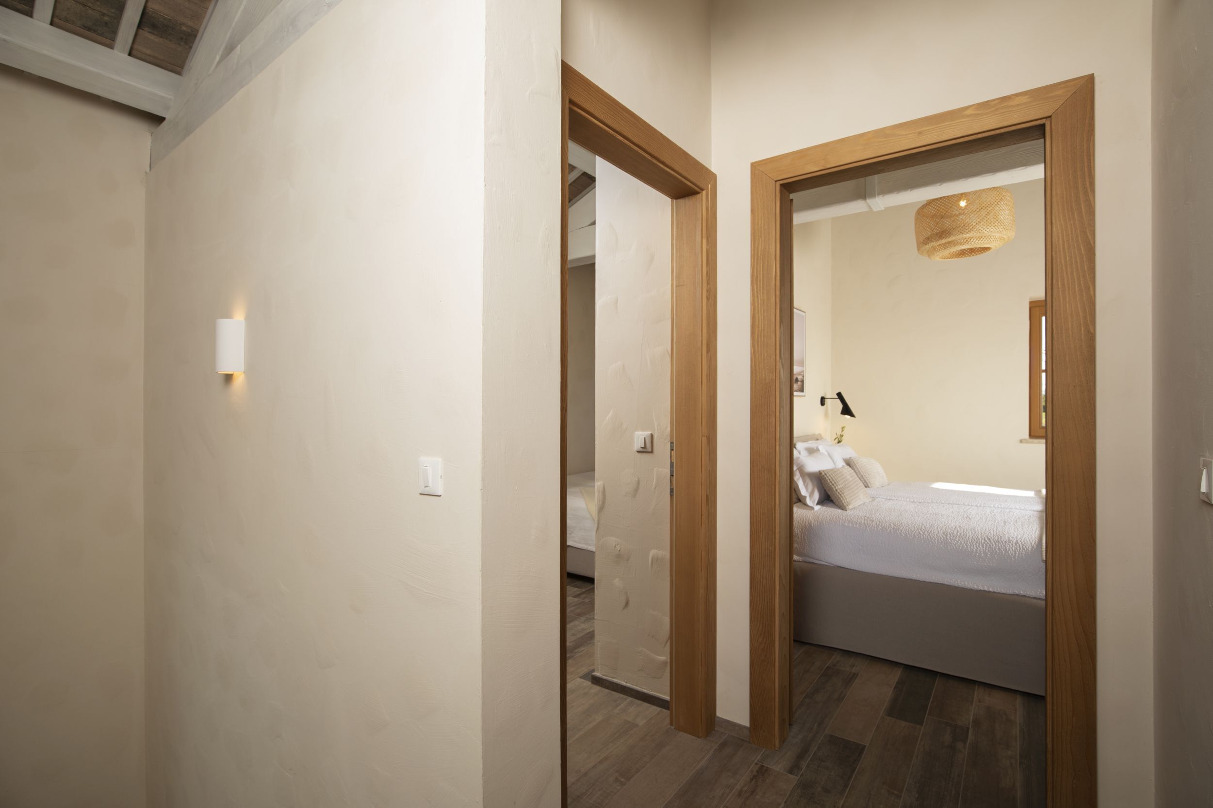 Una habitación de hotel moderna con una puerta de madera, una cama con ropa blanca y un baño visible al fondo.