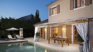 Zona junto a la piscina con piscina, sillas de descanso, una mesa y un porche cubierto.