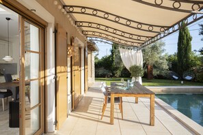 Un espace extérieur aménagé pour les repas, avec une piscine, une table et des chaises en bois, attenant à une maison dotée de grandes fenêtres et d’un patio couvert.