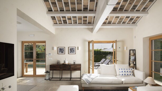 Amplia sala de estar con un sofá, una mesa de centro y un televisor. Hay un escritorio con una lámpara y una estantería. La habitación tiene ventanas grandes y vistas al exterior.