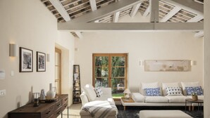 Una sala de estar moderna con sofá, reposapiés y una mesa de centro. Hay un escritorio de madera con cajones y una lámpara. La habitación tiene un ventanal con vistas a zonas verdes.