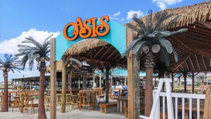 Un restaurante frente al mar llamado 'Crisis', con mesas y sillas de madera, palmeras y una estructura de techo de paja.