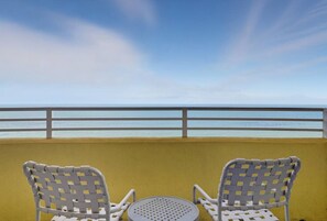 Two wicker chairs and a small round table on a balcony with a yellow wall and a railing, overlooking a beach.