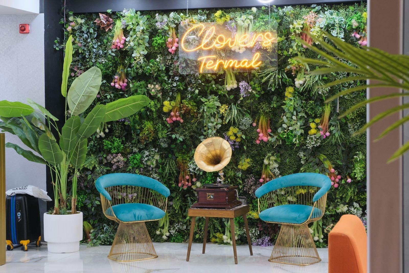 Una habitación con una pared decorada con diversas plantas y un letrero de neón que dice 'Lovers Terminal'. Hay dos sillas con cojines azules y un tocadiscos sobre una mesita.