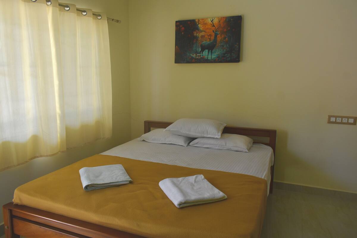 A neatly made bed with two folded towels, a painting on the wall, and a window with curtains.