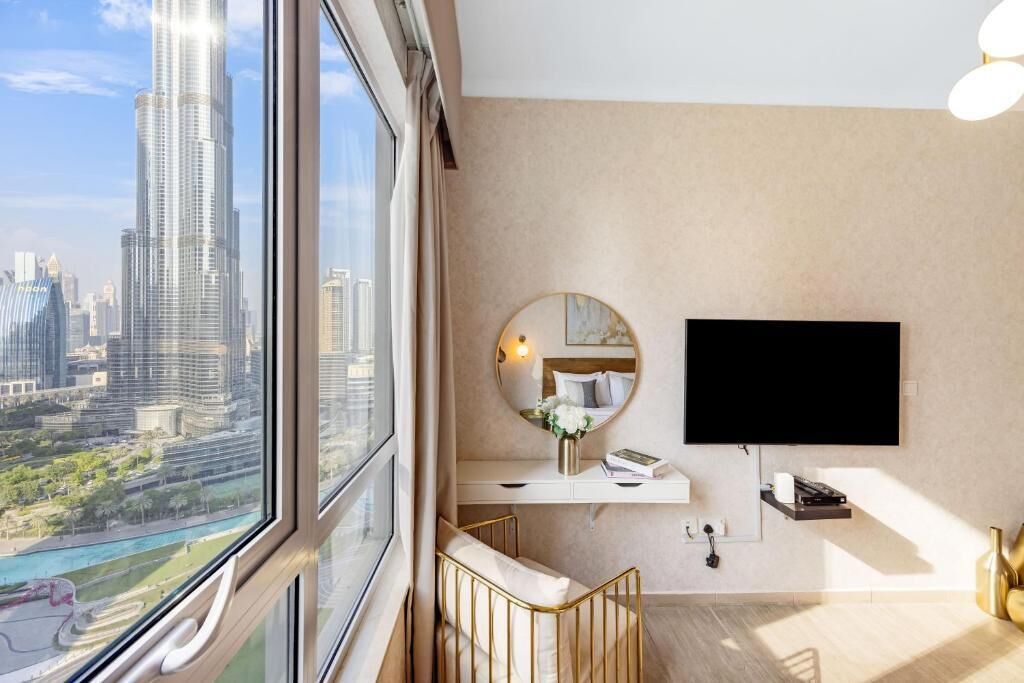 Une chambre d’hôtel moderne dotée d’une grande fenêtre donnant sur la ville, d’un téléviseur à écran plat fixé au mur, d’un miroir rond et d’un petit bureau agrémenté d’un vase de fleurs.