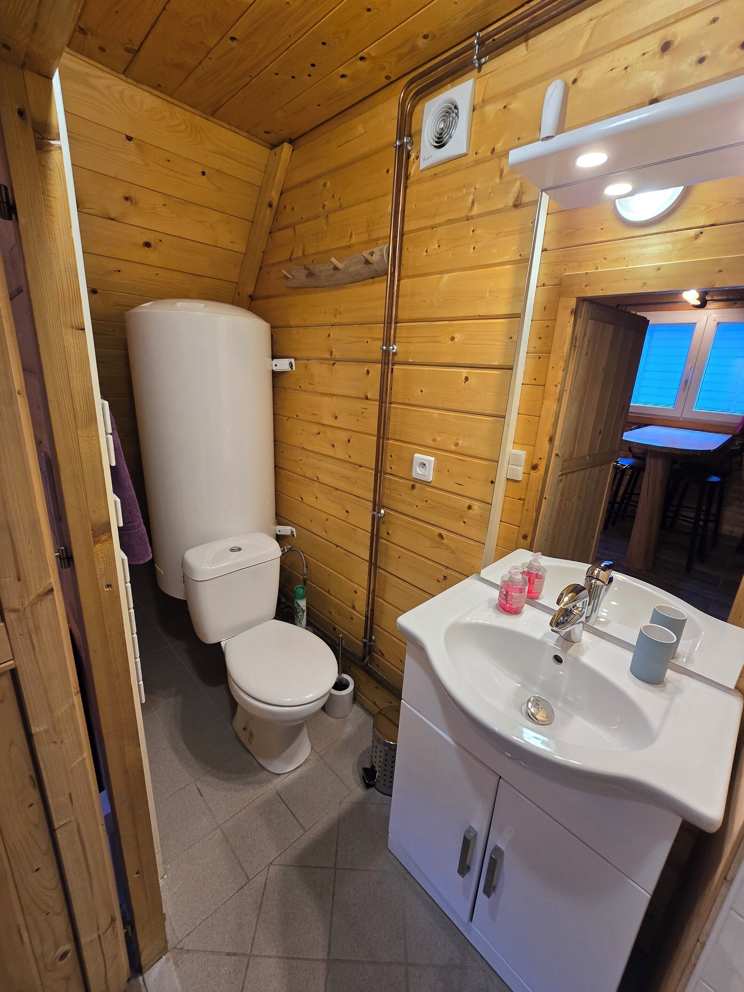 A compact wooden bathroom with a toilet, sink, and a window.