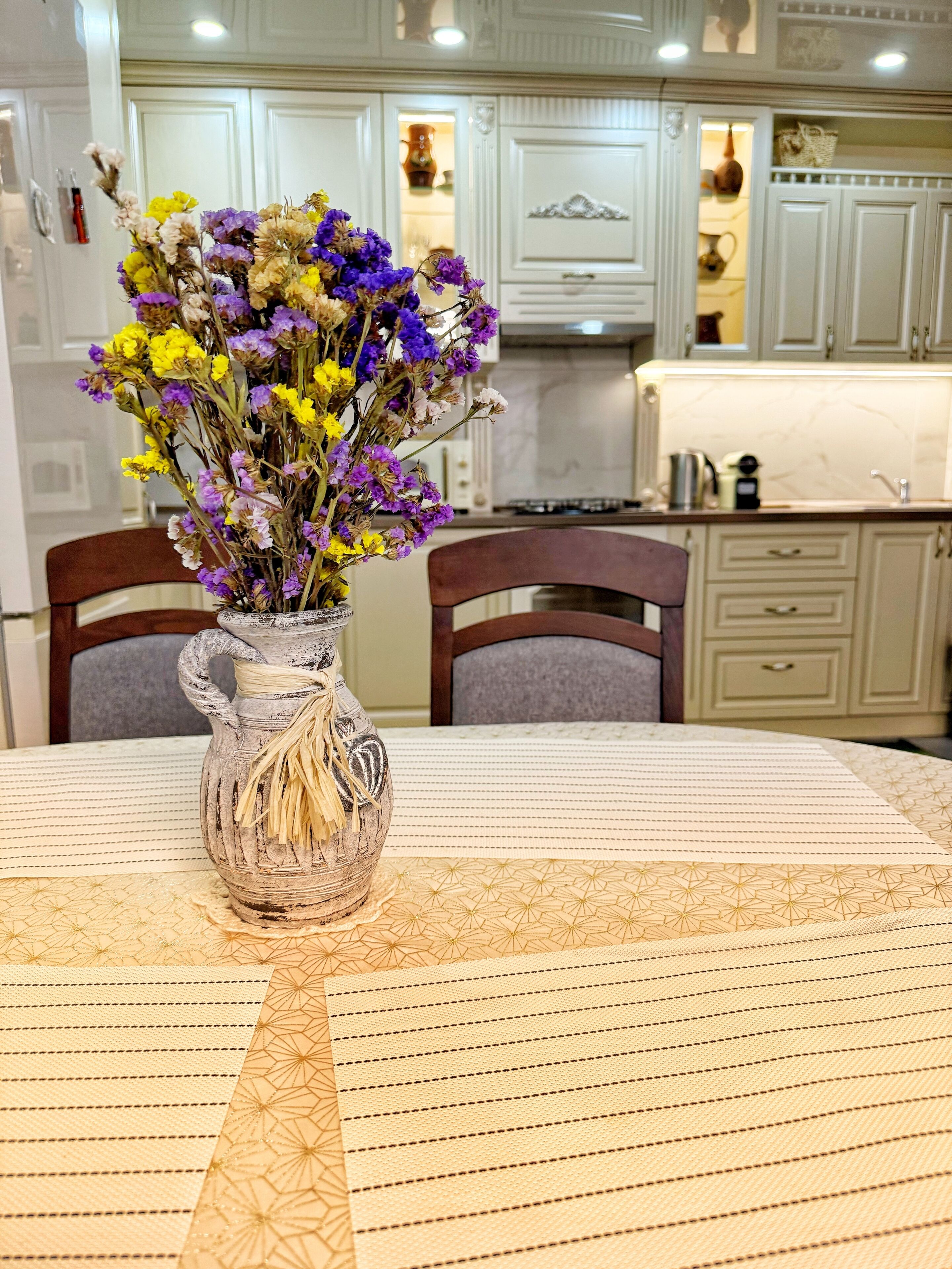 A dining table with a vase of flowers, chairs, and a kitchen in the background.