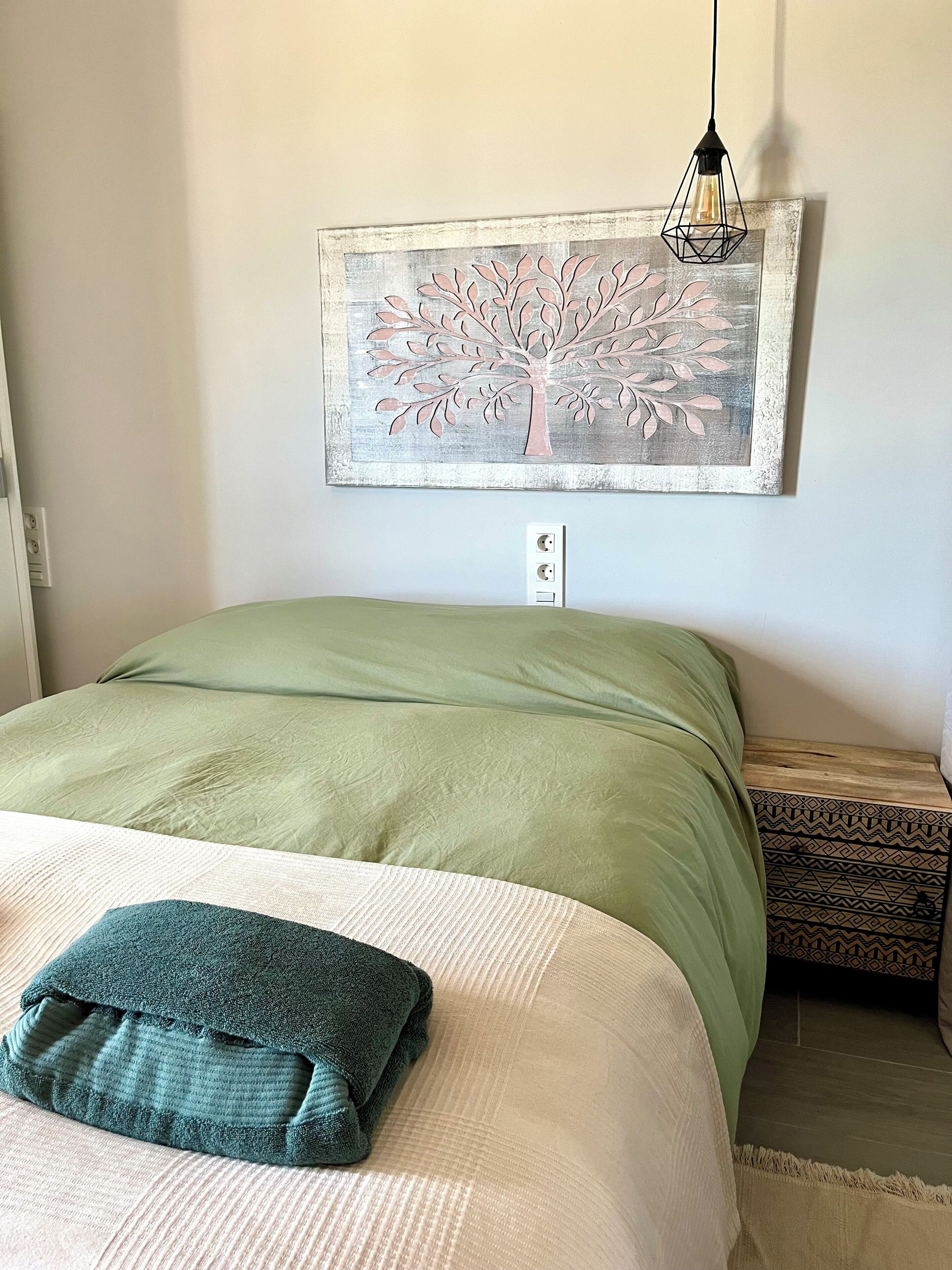 Un letto rifatto con copriletto verde e cuscino color acquamarina, un quadro di un albero appeso al muro e un comodino con un vassoio decorato.